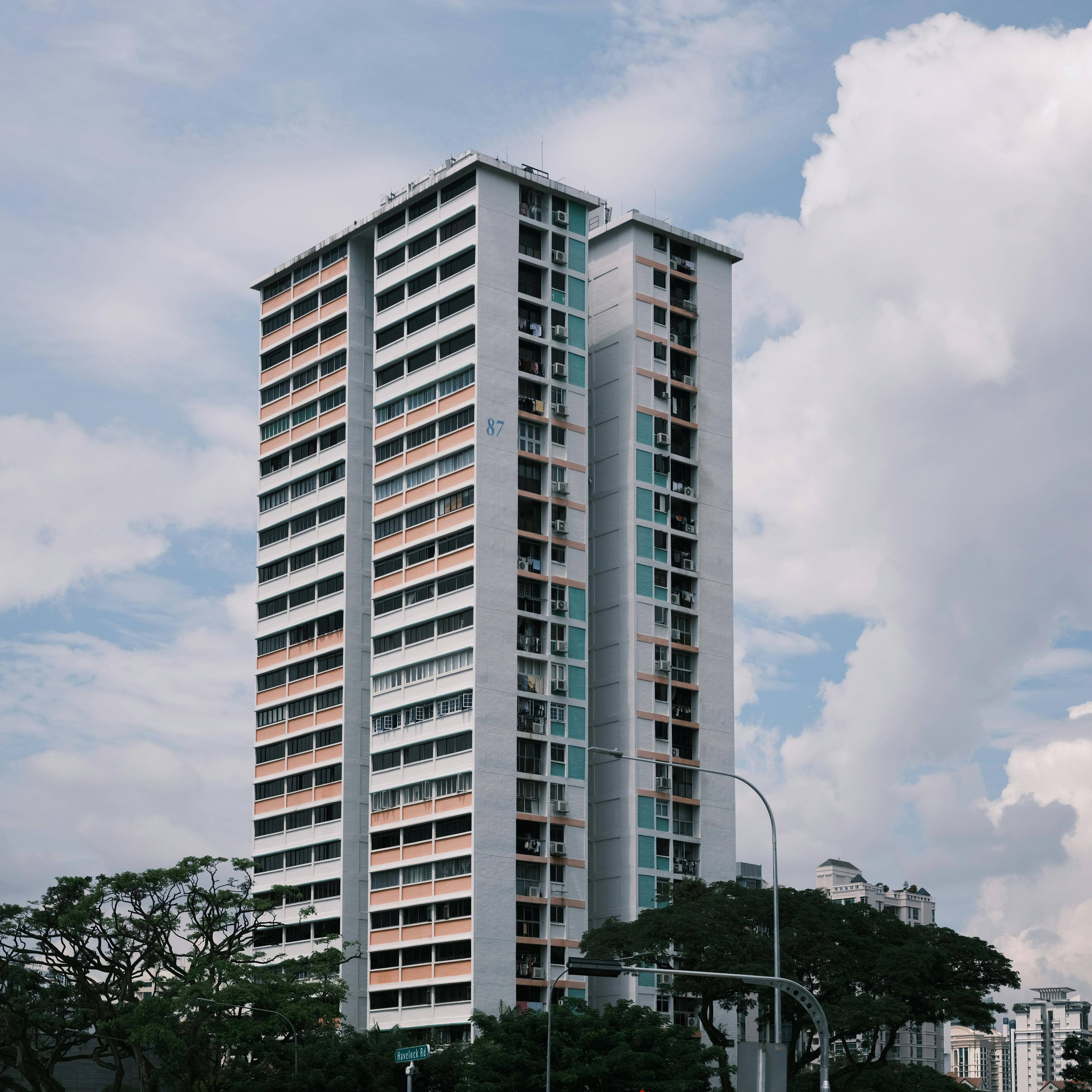 A tall white HDB point block with pastel panels against a cloudy sky