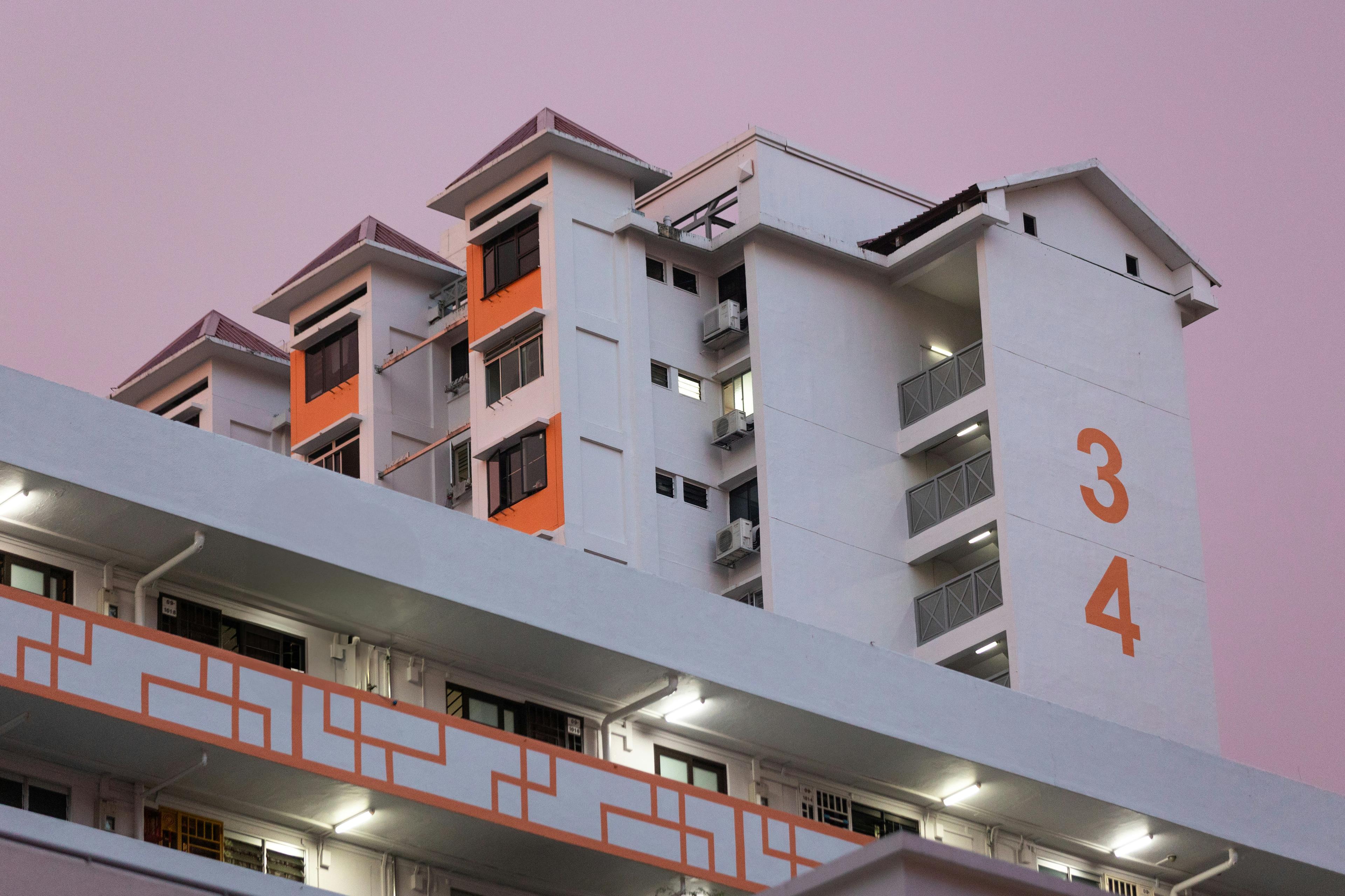The upper floors of a white HDB block with orange accents under a lilac evening sky